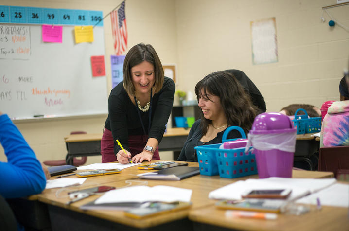 instructor leans over a desk to help a student in a classroom setting
