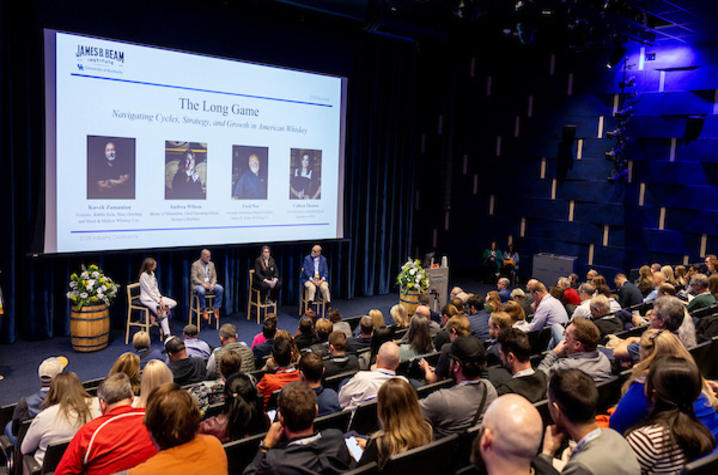 a panel of people sitting under a presentation screen at the front of an auditorium full of spectators