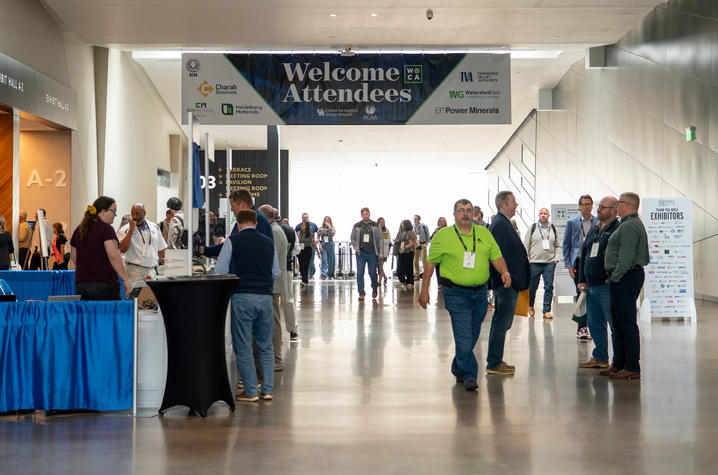 Wide view of a busy convention hall with attendees walking in and out beneath a large “Welcome Attendees” banner. Booths line the sides as people register, talk and move between sessions.