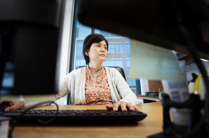 A researcher sits at a desk and works at a computer in an office, viewed from behind computer monitors and office equipment. Large windows in the background overlook another building.