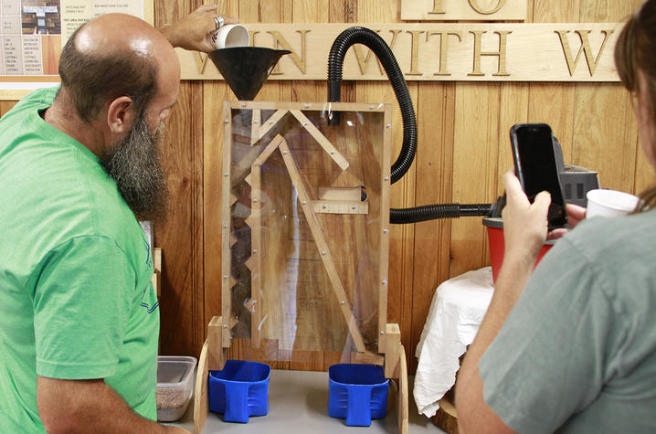 A person pours material from a cup into a funnel atop a transparent, wood-framed seed-sorting machine during a workshop demonstration.