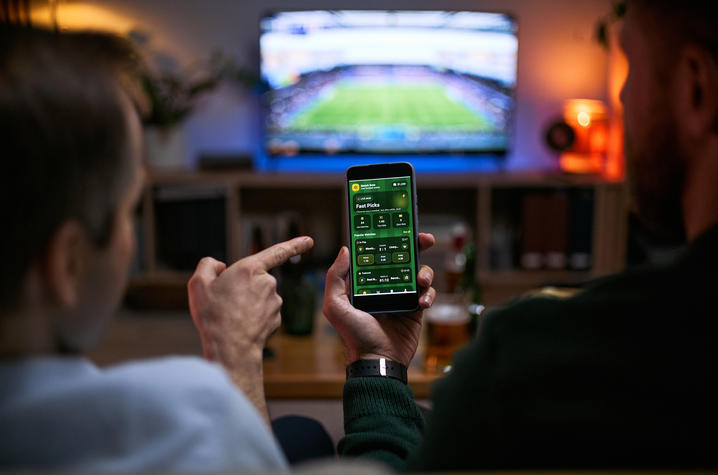 Two people sit in a living room watching a soccer match on a television while one person holds a smartphone displaying a sports betting app. 