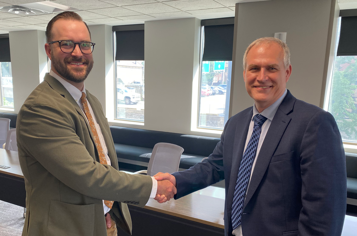 Two adults in business attire shake hands and smile in a conference room. Large windows line the room behind them, highlighting a professional partnership or collaborative agreement.