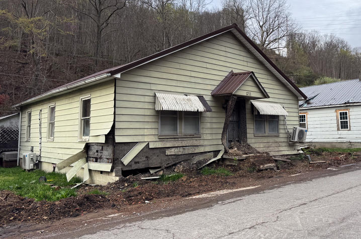 A small house shows significant damage, with siding torn away, a buckled foundation, and mud debris surrounding the property. The scene reflects the effects of a recent landslide or ground failure.