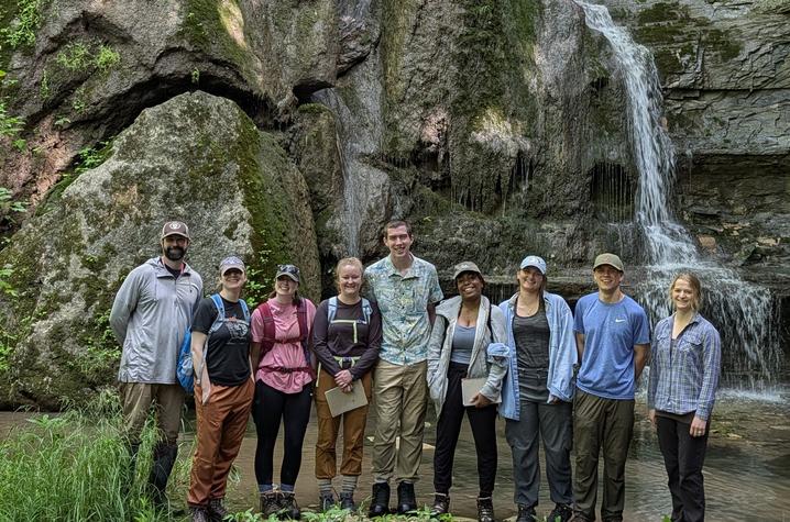 Nine people stand in front of a rocky waterfall and moss-covered cliff in a forested area.