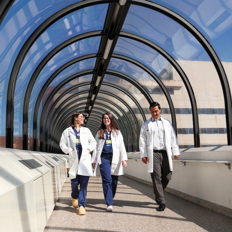Two people in white coats and blue scrubs and one person in a white coat, white shirt, and brown pants walking side by side in a well-lit pedestrian tunnel