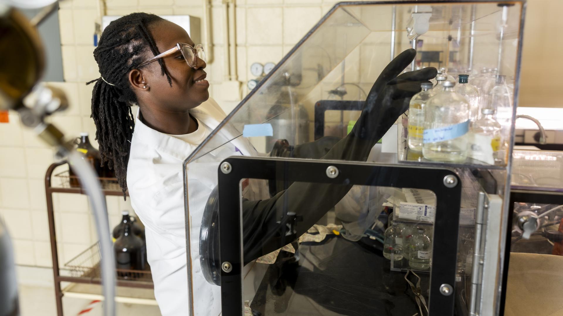 A student in a white coat with black gloves and eye protection reaching into a clear box demonstrating an experiment.