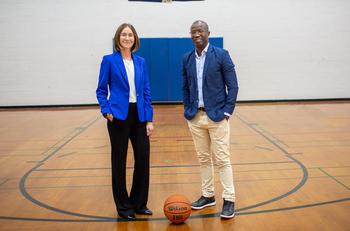  Two people stand on a gym basketball court beneath a hoop, with a basketball on the floor between them.