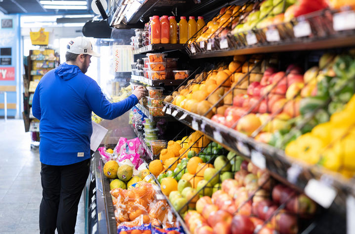 A shopper in a blue jacket and white cap browses a grocery store produce section, reaching toward packaged foods while standing beside shelves stocked with colorful fruits, vegetables and bottled juices.