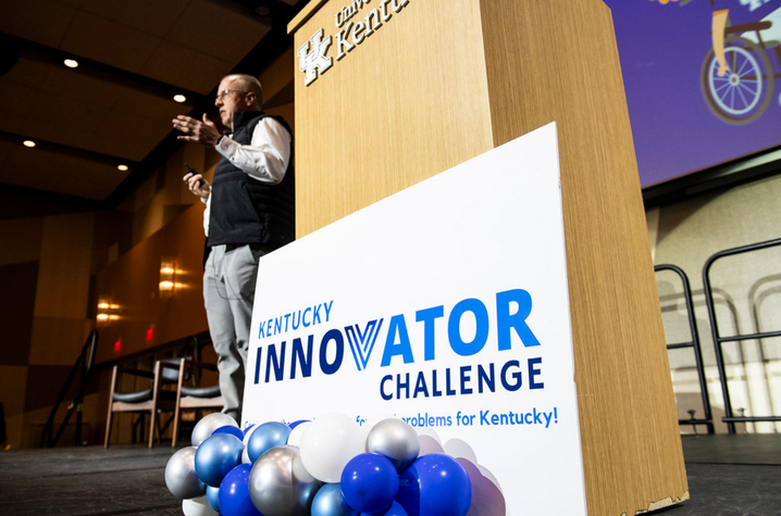 Presenter speaks beside a University of Kentucky podium at the Kentucky Innovator Challenge. Blue, white and silver balloons decorate the stage in a large conference hall.