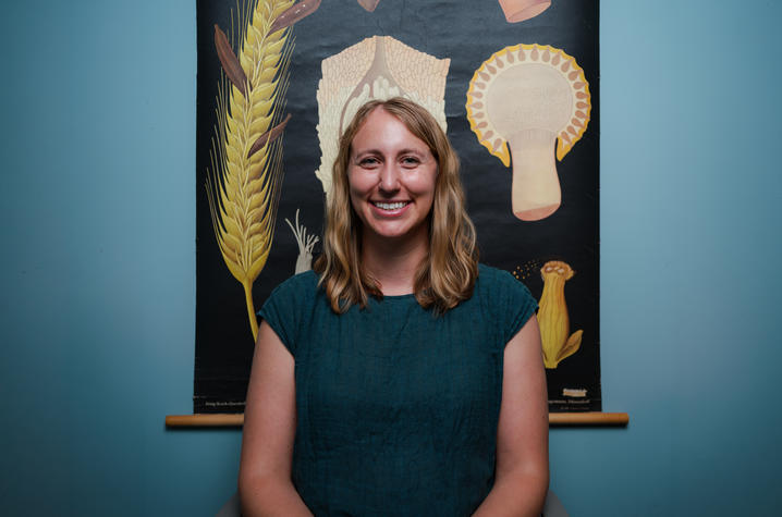 Person seated in a chair poses in front of a botanical wall chart featuring enlarged illustrations of fungal structures on a dark background.