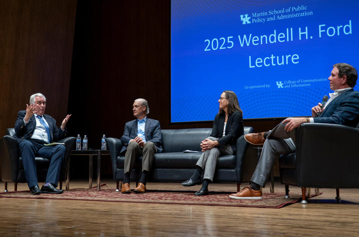 Four panelists sit on a stage during a public lecture and discussion at the University of Kentucky. One speaker gestures while talking from an armchair, while the other panelists listen from a sofa and adjacent chairs. A large screen behind them.