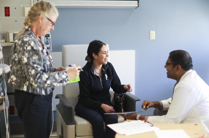 Three people gather in a clinical exam room, with one person seated and speaking with another seated person in a white coat while a third person stands nearby.