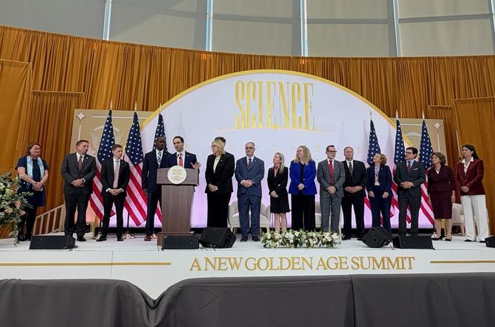 People stand on a stage beneath a large “A New Golden Age Summit” sign, with U.S. flags displayed behind them and a speaker at a podium addressing the audience.  
