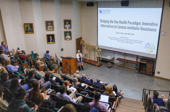 Presenter stands at the front of a lecture hall, speaking to an audience seated in tiered rows. A large projection screen displays a presentation titled “Bridging the One Health Paradigm.”