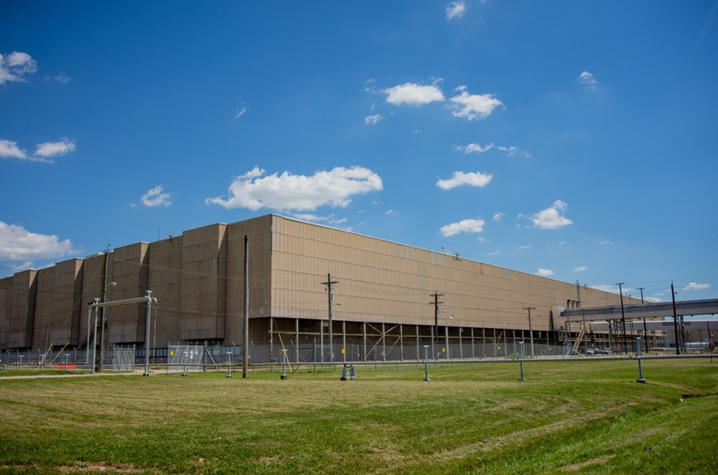 Large tan industrial building surrounded by fencing, utility poles and pipes, with a grassy field in the foreground beneath a bright blue sky with scattered clouds.