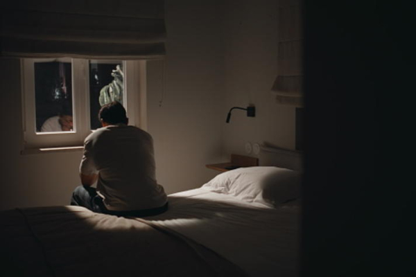 a person sits on the edge of a bed in a dark room while light comes in through a small window