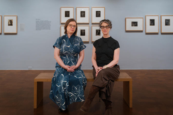 two people sit on a bench in an art gallery, facing away from photos arranged on a blue wall
