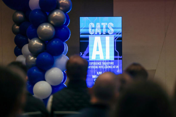 A digital screen reading "CATS AI" at the front of a room with an audience and a blue, white and silver balloon tower adjacent.