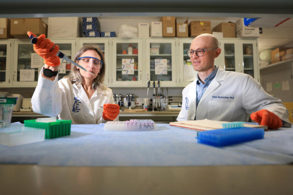Two researchers wearing lab coats, safety glasses and protective gloves work together at a laboratory bench. One researcher uses a pipette to transfer a sample while the other observes and takes notes, surrounded by scientific equipment and sample trays.