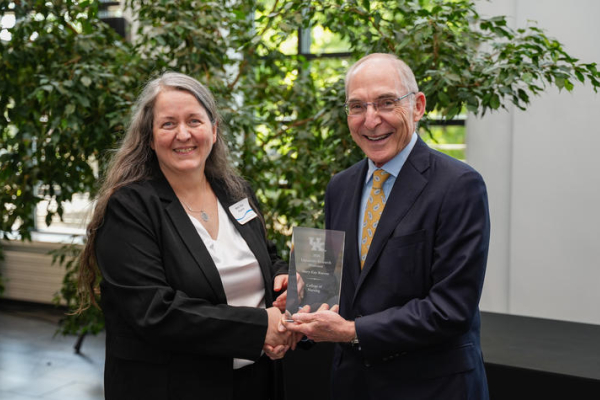 Two people shake hands while holding a glass award during a recognition ceremony. They stand in a bright indoor space with large leafy plants behind them, celebrating an academic achievement.