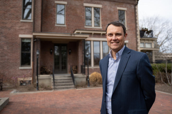 A person in a blue blazer stands in a brick courtyard in front of a large historic red-brick building with tall windows and a covered entrance. The building is slightly out of focus in the background.