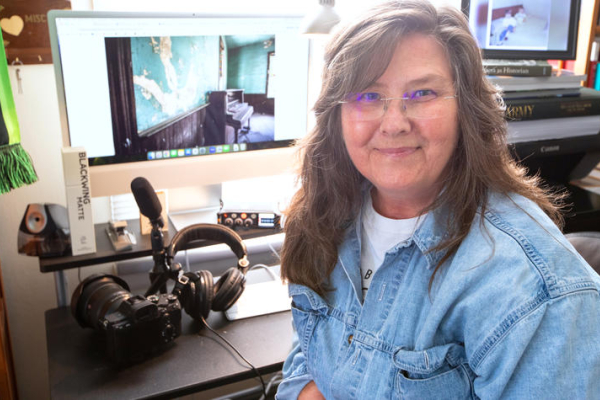 Kopana Terry sits at an office workstation with a camera, microphone, headphones and computer displays showing images of historic interiors. The workspace reflects efforts to document, preserve and study cultural heritage sites.