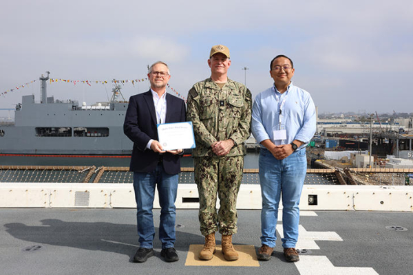 three men, one in a blazer holding a certificate, one in Navy uniform and one in a button-down shirt, stand together on the deck of a ship with another Navy ship behind them