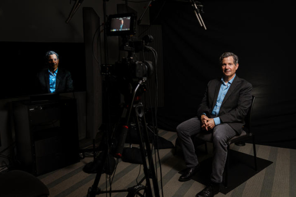 A person sits in a recording studio during a video interview. A camera, lighting equipment and microphones are visible in the foreground, capturing the subject against a dark backdrop while a monitor displays the live video feed.