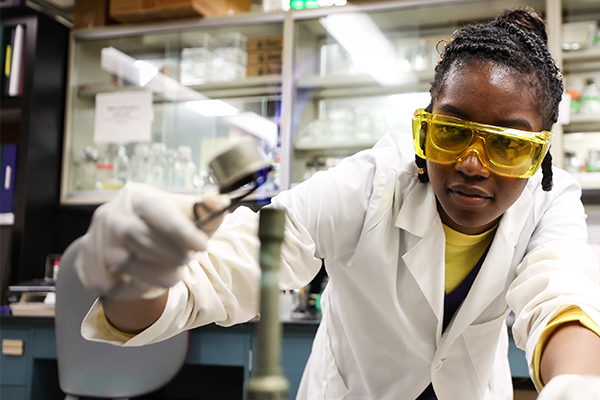 a person wearing a lab coat, goggles and gloves lights a Bunsen burner in a laboratory