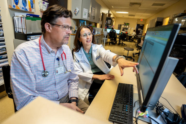 Two health care providers review information on a computer workstation in a hospital clinic. One points to data on the screen while discussing results, illustrating collaboration in patient care and clinical decision-making.