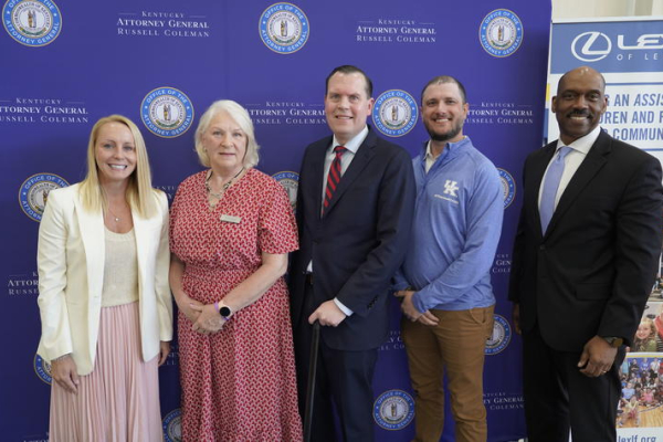Five people stand in front of a blue Commonwealth of Kentucky-branded backdrop during an event, with a vertical display banner visible at the right side of the photo.