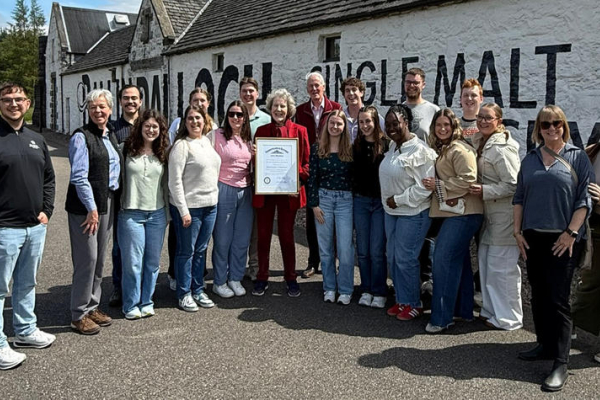 A group of about 20 people stand outdoors in front of a white stone building with large black painted lettering on the wall. One person in the center holds a framed certificate while the group gathers around for a photo.