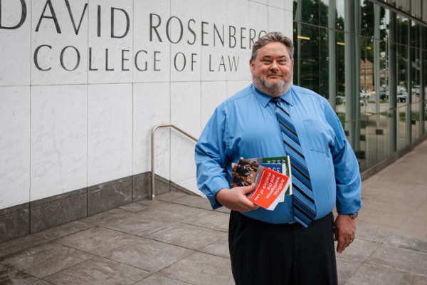 A person stands outside the Rosenberg College of Law building, holding several books.
