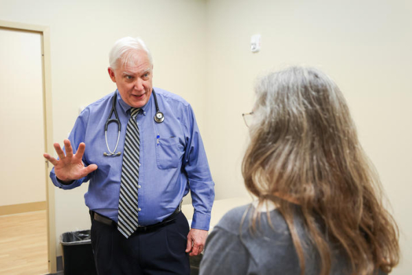 A physician wearing a blue shirt, tie and stethoscope gestures while speaking with a seated patient during a clinical visit.
