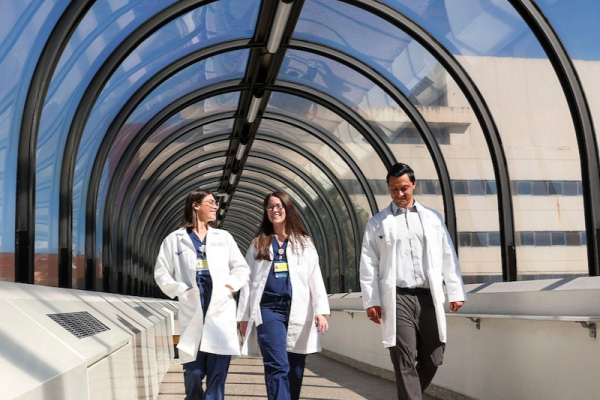 Two people in white coats and blue scrubs and one person in a white coat, white shirt, and brown pants walking side by side in a well-lit pedestrian tunnel