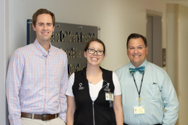 Three healthcare team members stand together in a hospital hallway in front of a sign for a pediatric oncology clinic. One person wears a plaid button-down shirt, another wears a black hospital vest with an ID badge, and the third wears a light blue shirt
