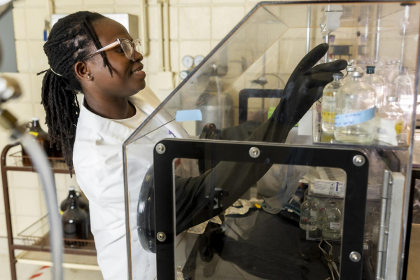 A student in a white coat with black gloves and eye protection reaching into a clear box demonstrating an experiment.