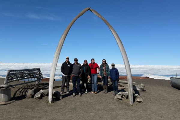 Six people stand beneath a large arch formed by two whale bones on a dark shoreline, with wooden boats and a vast expanse of sea ice beneath a clear blue sky in the background.