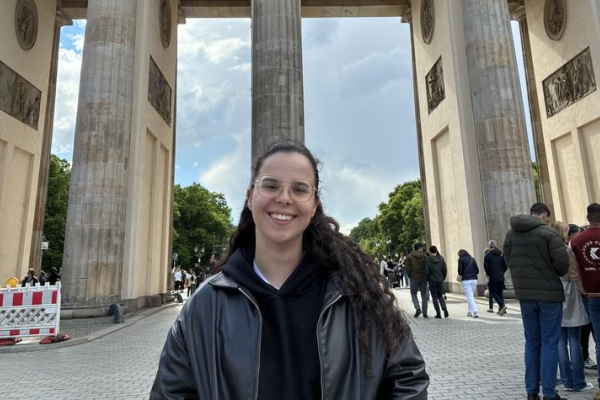 Person standing in front of the Brandenburg Gate in Berlin, Germany, with the landmark’s columns and the Quadriga sculpture visible overhead. Pedestrians and an overcast sky appear in the background.