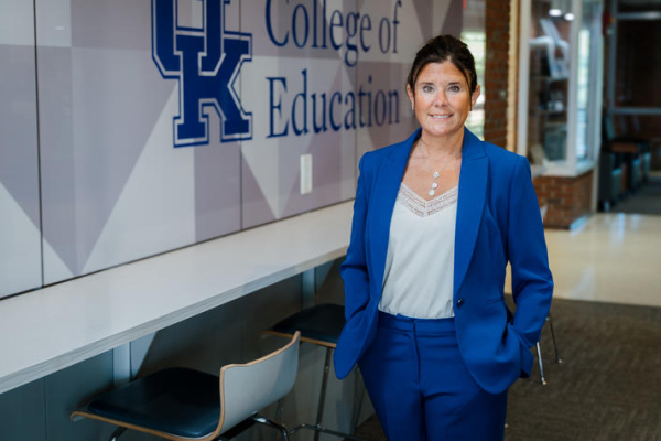 A person in a blue suit stands in a hallway beside a wall displaying the University of Kentucky logo and the words “College of Education.” A counter with stools lines the wall, and the hallway extends into the background.