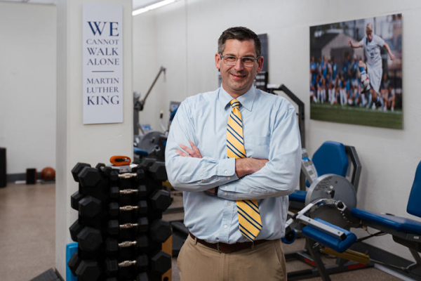 Person stands with arms crossed in a biomechanics lab containing dumbbells, exercise equipment and wall displays, including a sign reading, “We cannot walk alone.”