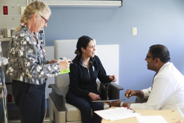 Three people gather in a clinical exam room, with one person seated and speaking with another seated person in a white coat while a third person stands nearby.