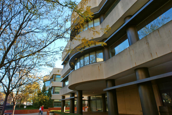 Exterior view of a multi-story campus building with curved concrete balconies, large windows and cylindrical support columns. Bare tree branches frame the left side of the image, while a sidewalk and landscaped areas surround the building.