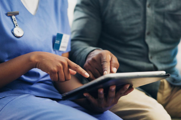 Health care worker in blue scrubs and a person in a gray-green shirt review information together on a tablet.