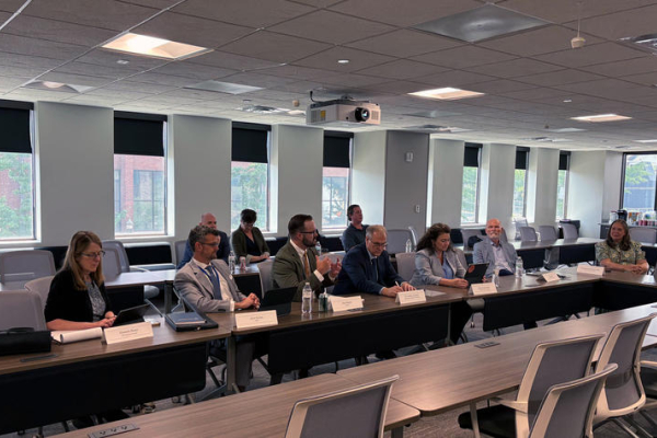 A group of people seated around U-shaped conference tables during a meeting in a large room with windows, laptops, notebooks, water bottles and name placards arranged on the tables.