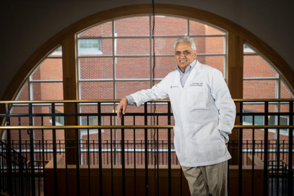 Person in a white lab coat stands beside a railing in front of a large arched window.