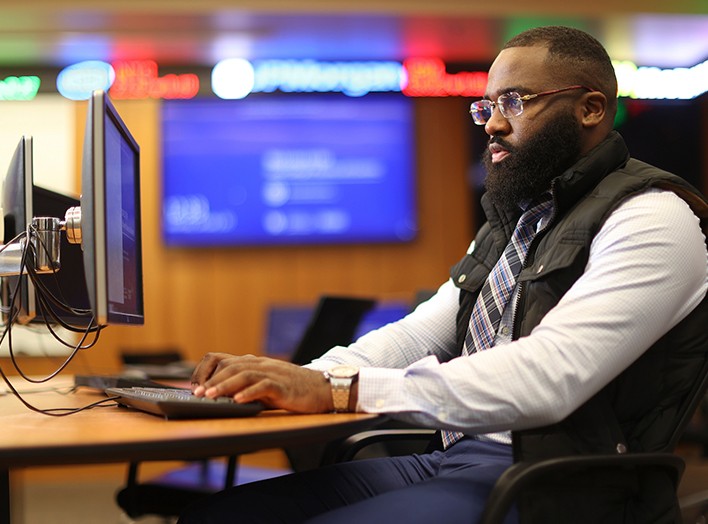 A man with glasses and a beard, wearing a white shirt, tie, and a vest, sits at a desk working on a computer. Behind him, the room is illuminated with neon signs and screens, suggesting a modern office or newsroom setting. He appears focused.