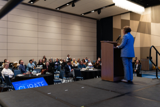 Ilhem Messaoudi speaks at a podium on stage while event attendees look on from tables