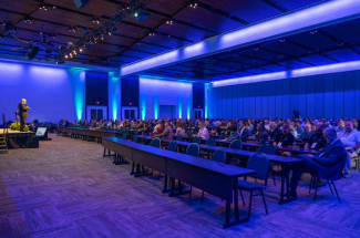 Large conference room filled with seated attendees facing a speaker at the front. Blue and green lighting illuminates the space, with rows of tables and a stage presentation underway.